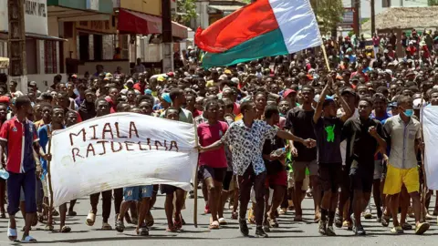 AFP via Getty Images Dozens of protesters can be seen marching down a road. At the front are people holding a banner which says 'Rajoelina resign' in Malagasy. A Madagascan flag can also be seen. 