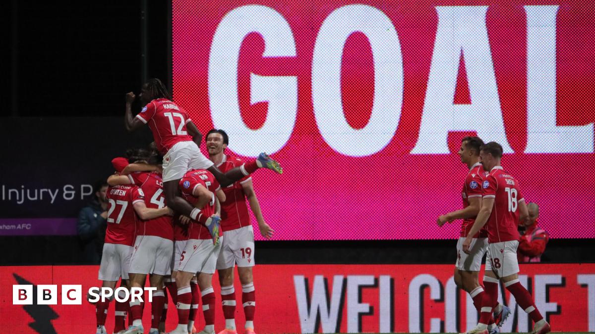 Wrexham players celebrate in a huddle in front of a large scoreboard with the words 'Goal'