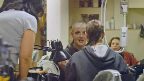 Bauer-Griffin via Getty Images Britney Spears is seen seated in a hair salon with her head half shaved. She is smiling looking at herself in a mirror as an attendant hovers over her shoulder. 