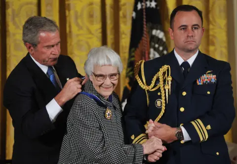 Getty Images President George W. Bush in dark suit and blue tie, putting the  Presidential Medal of Freedom around Harper Lee's neck. Harper smiling, wearing glasses, dressed in black and white dog tooth outfit, holding hand of man in uniform