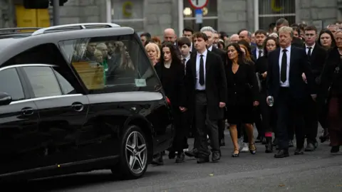 PA  Media A hearse with a coffin inside is moving along a road with a procession of people, dressed in black, behind it.