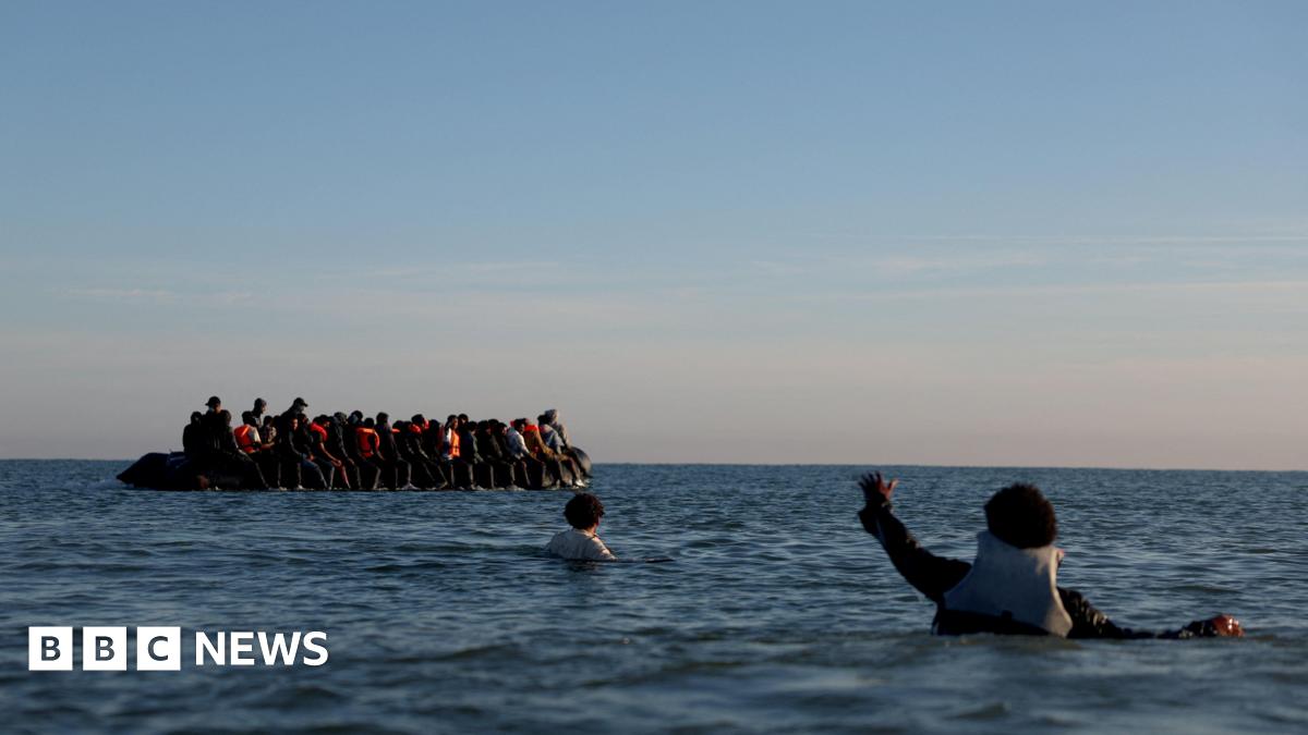 A group of migrants on an inflatable dinghy leave the coast of northern France in an attempt to cross the English Channel to reach Britain, from a beach in northern France. In the foreground of the picture there are two people who are in the water. One is holding out their hand to towards the dinghy