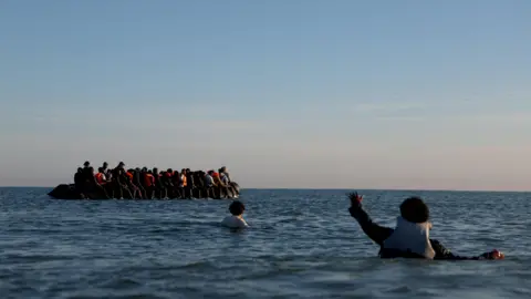 Reuters A group of migrants on an inflatable dinghy leave the coast of northern France in an attempt to cross the English Channel to reach Britain, from a beach in northern France. In the foreground of the picture there are two people who are in the water. One is holding out their hand to towards the dinghy