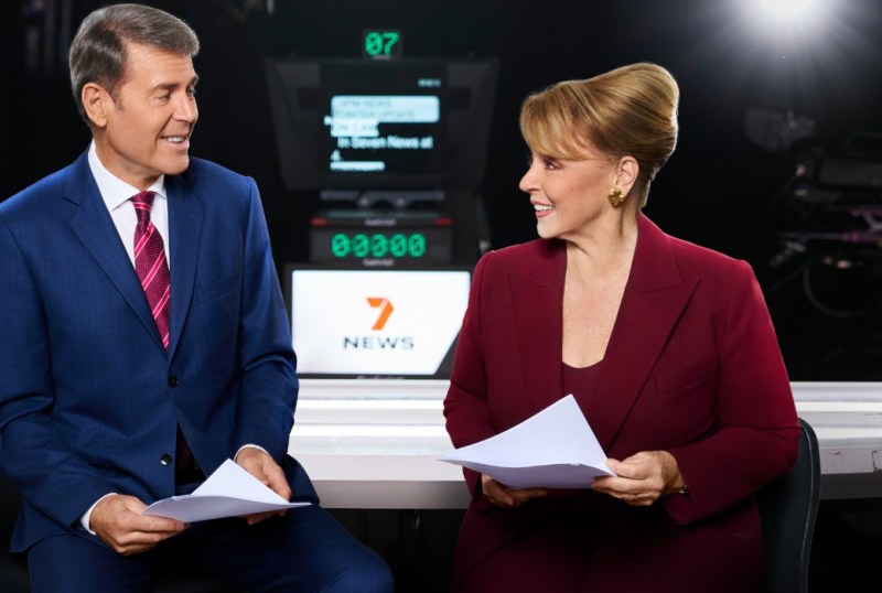 Two news presenters smile at each other while holding scripts in a television studio, with a '7 News' logo and teleprompter in the background. The male presenter is dressed in a blue suit with a pink tie, while the female presenter wears a burgundy outfit.
