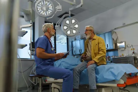 Getty Images Doctor dressed in blue scrubs talks to older patient dressed in jeans and yellow jacket sat on a hospital bed.