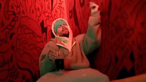 Justin Bieber/Instagram Justin Bieber holding a pint glass half-full of a dark-coloured liquid. He is sitting on an upholstered yellow bench and leaning against a red wall decorated with black line-drawings. He appears to be singing with his eyes closed. 