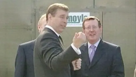 Prince Andrew has short brown hair and is gesturing towards the camera. Mr Trimble has short hair combed to one side. He is wearing glasses, a dark suit, white shirt and purple tie.