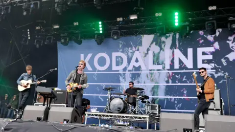 Kodaline play on stage. There is a banner behind them which says 'Kodaline'. 