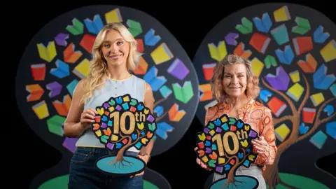BBC Ali and Cecilia are both holding Book Week symbols, which are in the shape of a tree with multi-coloured leaves and the word 10th in the centre. Ali has long blonde hair and is wearing a blue sleeveless top. Cecelia has shoulder length hair and is wearing an an orange patterned blouse.