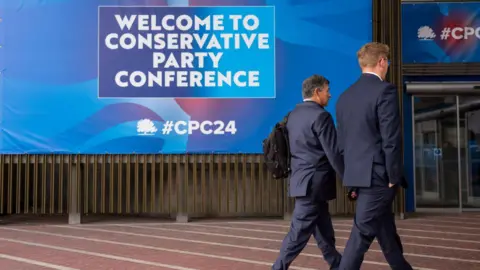 Getty Images Two men in suits walk past a huge poster at the Conservative Party Conference in Manchester. The poster reads: welcome to conservative party conference.