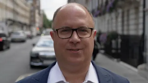 Adam Hug standing in a street in Westminster. He is a white man with tortoiseshell glasses and is wearing an open white shirt and a blue blazer