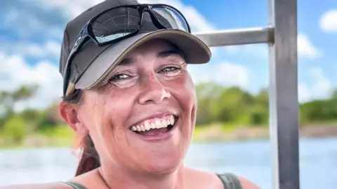 Isobel Wright Ms Wright looking to be enjoying a sunny day outdoors, smiling warmly with a cap and sunglasses perched on top of her head. Behind her is a scenic landscape with a body of water, lush greenery, and a partly cloudy blue sky.