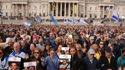 Reuters A large crowd of men and women gathered in Trafalgar Square. Many hold or a draped in blue and white Israeli flags, and some Union Jacks. Others hold posters of some of the hostages, which features a picture of them, their ages and their names, printed in red, capital letters, in both English and Israeli. In the background more people can be seen lining the steps around the National Gallery.