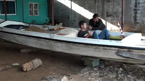Gustavo Ocando Alex A man sits inside a small boat propped up on dry land while another leans on the boat to speak to him
