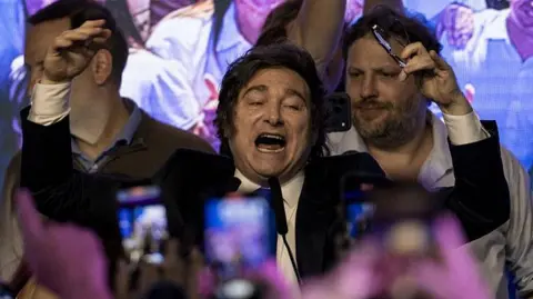 Getty Images Javier Milei wearing a suit, surrounded by people and with smartphones in the foreground filming him, throws his hands in the air with his mouth open