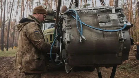 Getty Images A man in military garb, beanie and sunglasses, with a Ukraine flag badge on his jacket sleeve, tends to a large piece of machinery in the middle of the woods