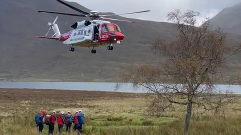 Skye Mountain Rescue Team A red and white helicopter landing on a green strip of grass with mountains and a loch behind.