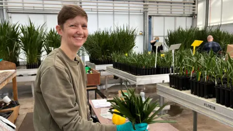 BBC Woman with brown hair smiling while holding plants inside a building filled with plants. She is wearing a green top and blue gloves.