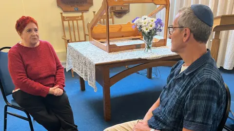Jennifer Clements wearing black trousers and a knitted bright red jumper which matches her hair, which is cut very short. She is sitting on a blue chair opposite a male member of the Jewish congregation who is wearing a blue and black checked shirt. 