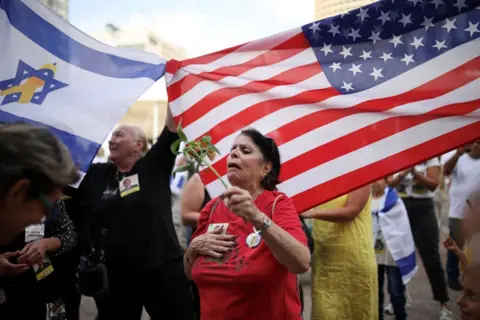 Reuters Woman holding flower seems overcome with emotion, as crowds behind wave USA and Israel flags