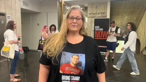 BBC A woman wearing a T-shirt that displays a picture of a younger man with the words "bring Tamir home". She is looking at the camera, and people are walking past her in the background