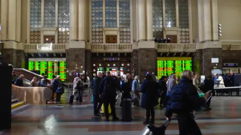 Kat Werner/BBC People rush through Kyiv central station, in front of large departure boards announcing the trains leaving in green text