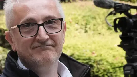 PA Media Moussa Kadri is seen leaving court. He is wearing a shirt, navy jacket and glasses. A press camera can be seen on a tripod behind him.