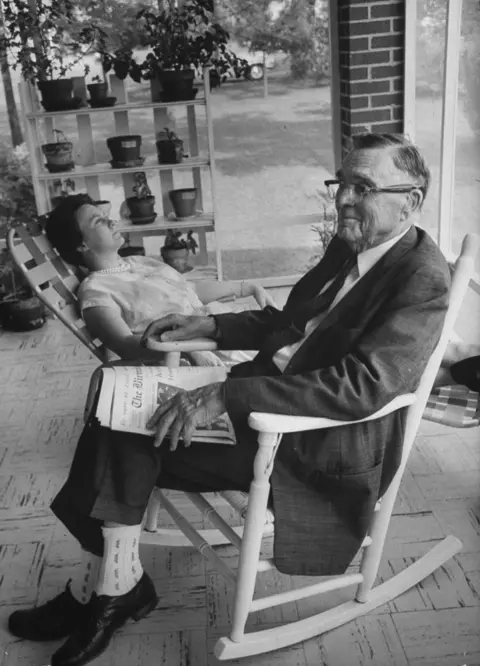 Getty Images Harper Lee  sitting outside on a reclining chair in short sleeved dress looking upwards, next to her father in dark suit and glasses, with a newspaper on his lap. Plants on shelf in the background