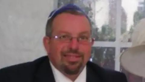 Facebook A man smiles at the camera. He is wearing a blue kippah on his head and glasses, and has a goatee. He is wearing a black suit jacket and a white shirt. Behind him is a blurred window. 