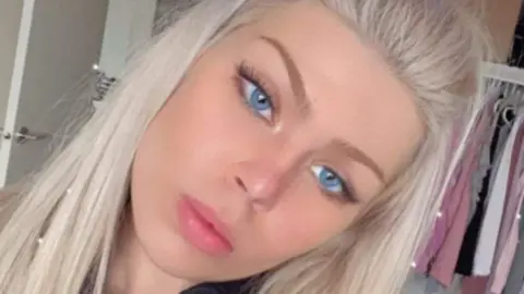 Police Scotland Young woman with long blonde hair and blue eyes, looking at the camera, with clothes hanging up behind her, and an open door.