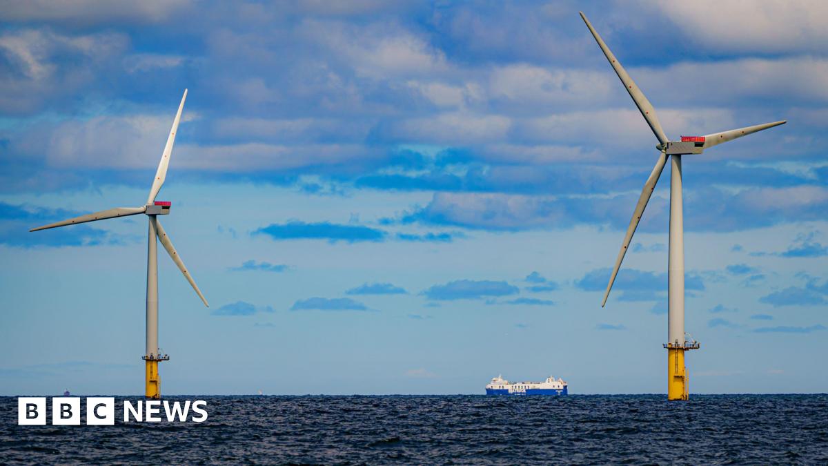 A ship passing between two offshore wind turbines on a dark blue sea under a light blue sky with grey and white clouds.