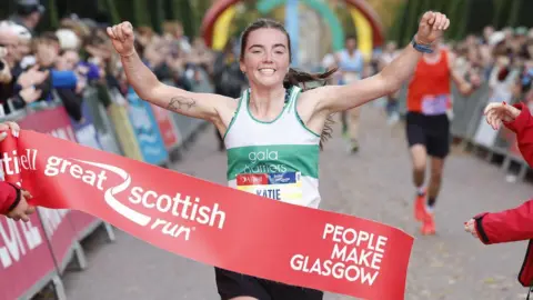 Great Scottish Run a runner wearing a number bib that reads 'Katie' and a white and green vest that says 'Gala Harriers', crosses the finish line of the Great Scottish Run with arms raised. The finish tape reads "People Make Glasgow".