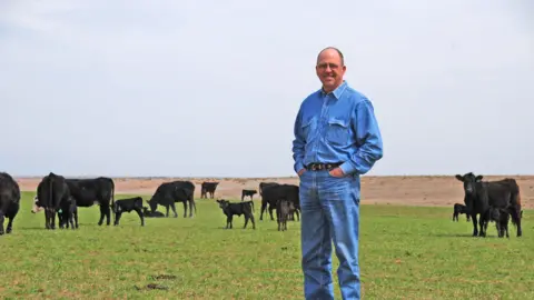 Mike Callicrate A man wearing denim stands on a grassy plot of land with his hands in his pockets. Several cows graze the land in the background. 