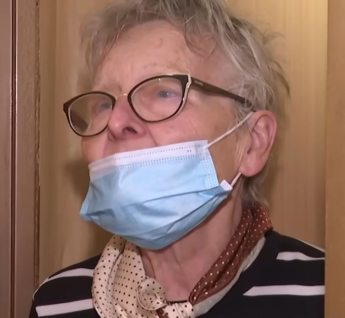 Elderly woman wearing glasses and face mask, speaking indoors about teen missing 27 years found living in parents’ house.