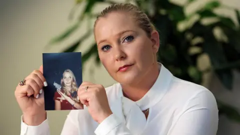 Getty Images Virginia Giuffre sits in a white blouse holding a picture of herself as a teenager
