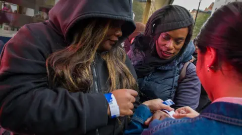 Two people wearing hoodies and jackets get wristbands put on them