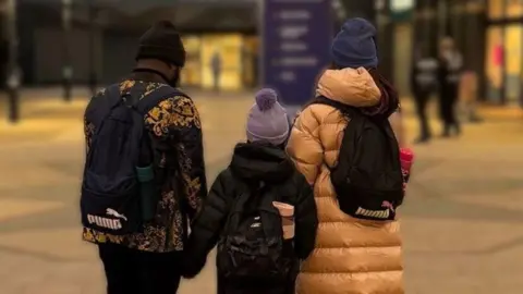 Supplied Photo of the backs of a man and two children next to him on the right. They are all facing away at the camera. They are outside and all wearing black backpacks and beanies. The man is wearing a black beanie and a black coat with white patterns. The smallest girl in the middle is wearing a purple beanie, a black coat and has a pink water bottle in her bag. The tallest girl is wearing a pink bag and a black backpack with the words "Puma" and is wearing a dark blue beanie. They are in a city centre space in the dark with the background blurred. 