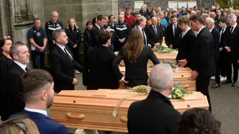 PA Media Three coffins in a row outside a church. There are flowers on each of them and people all around them, dressed in black.