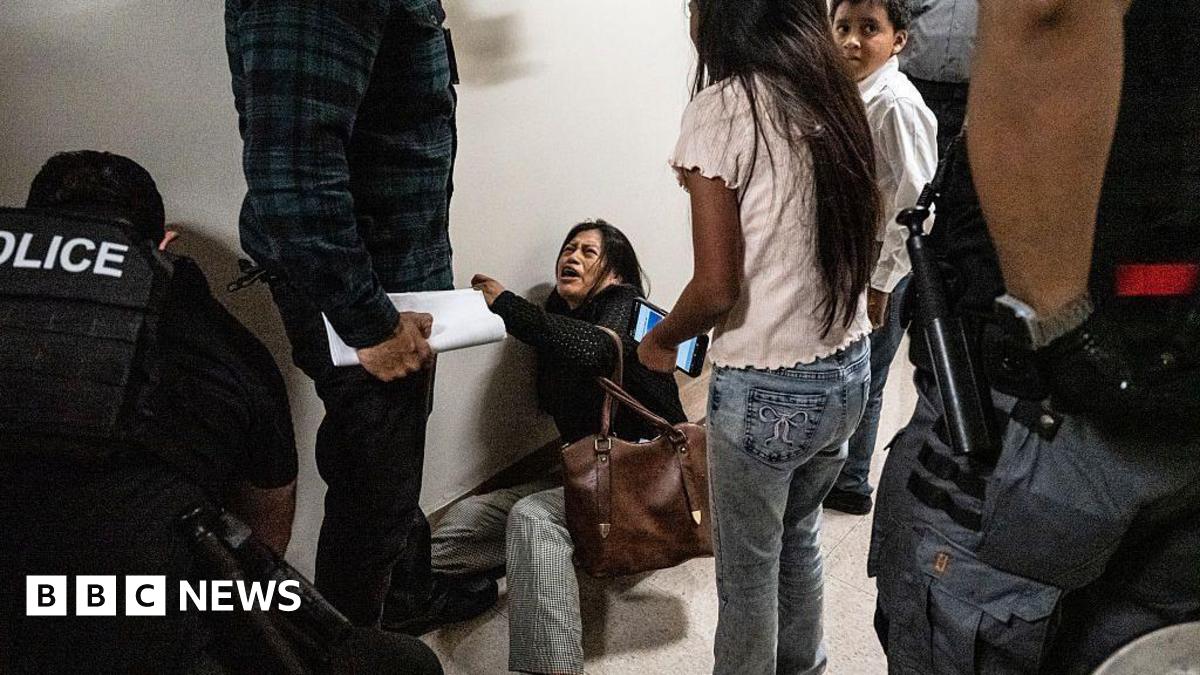 A woman lying on the floor cries. She is surrounded by officers wearing vests labelled police, and her two young children