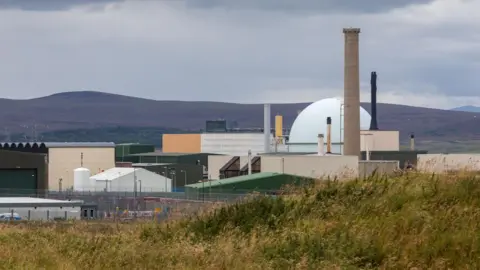 Getty Images The power station is a complex of buildings and structures, including metal sheds with peaked roofs, a chimney and a large sphere. In the foreground is an area of scrubby land, and in the background low hills.