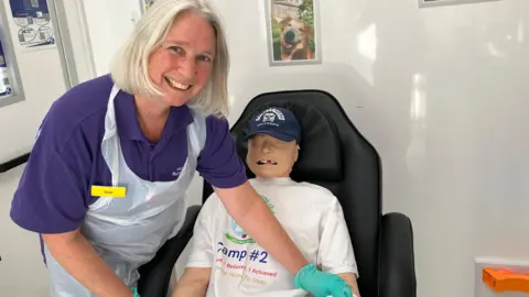 Henry Godfrey-Evans/BBC A woman with short blonde hair in a purple polo shirt and a white plastic apron smiling while leaning over a dummy which is placed sitting down on a black armchair.
