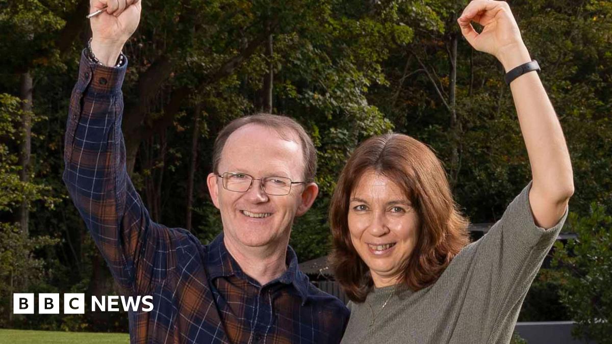 Richard and Yannick Baker stand in front of the house next to the swimming pool. Behind them are big trees. Richard is wearing a checked red and navy shirt with blue skinny jeans. He is wearing glasses and has short hair. Yannick is wearing black skinny jeans and is wearing a taupe shirt. She has long brown hair.