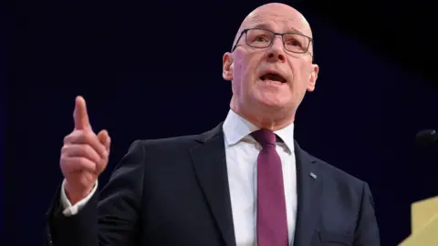 Getty Images John Swinney standing at a podium. He is wearing a dark suit over a white shirt with a purple tie. He is standing against a black backdrop with his right index finger pointing in the air.