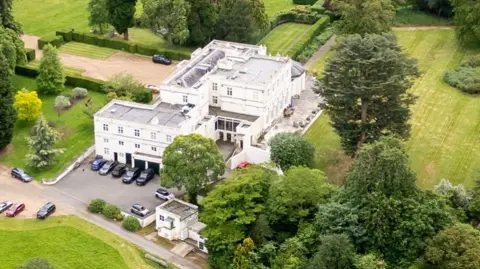 Shutterstock An aerial photograph of Royal Lodge, Prince Andrew's residence in Windsor, shows a large white building with green fields and trees surrounding it and a variety of cars parked outside it. 