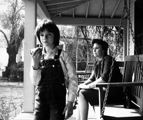 Getty Images Actress Mary Badham, short pudding bowl styledark hair eating an apple & dressed in dungarees and Harper Lee  seated on swing bench in porch on the set of the film "To Kill A Mockingbird"