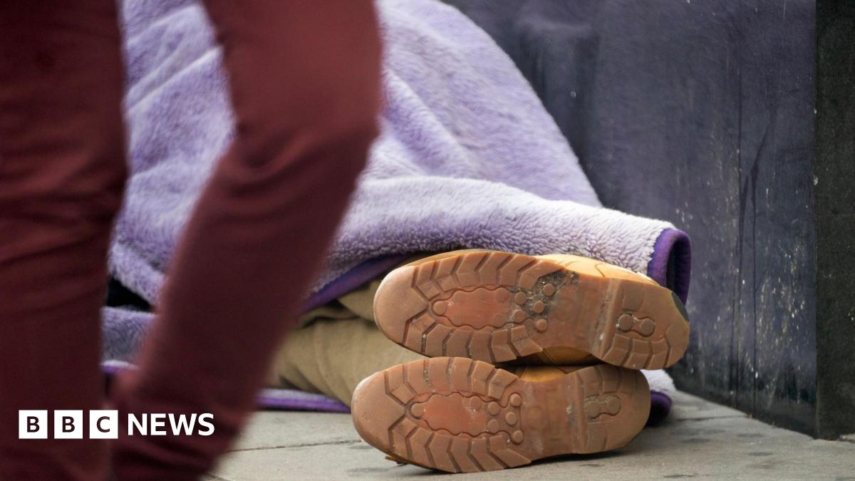 A person lies on the pavement wrapped in a purple blanket, with only their boots visible, as someone walks past in the foreground.