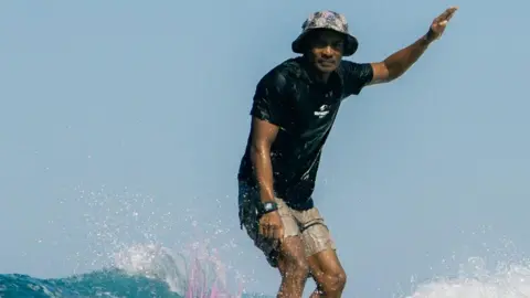 Supplied Damar wearing a bucket hat, black t-shirt and board shorts surfing on a wave, with water splashing around him. His hand is pointed in the air for balance, and he is looking intently into the distance.