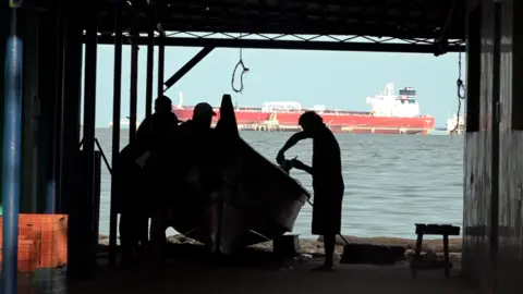 Gustavo Ocando Alex Four silhouetted men next to a boat in a covered space facing the sea
