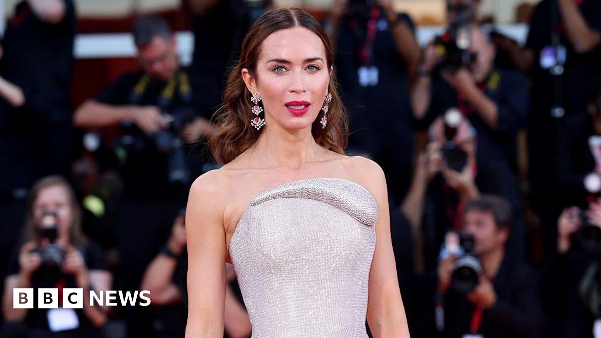 Emily Blunt stands on a red carpet. She is wearing a silver dress with earrings.