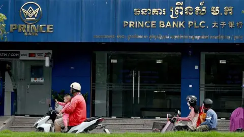 AFP via Getty Images Motorists ride past a branch of the Prince Bank in Phnom Penh on October 15, 2025.